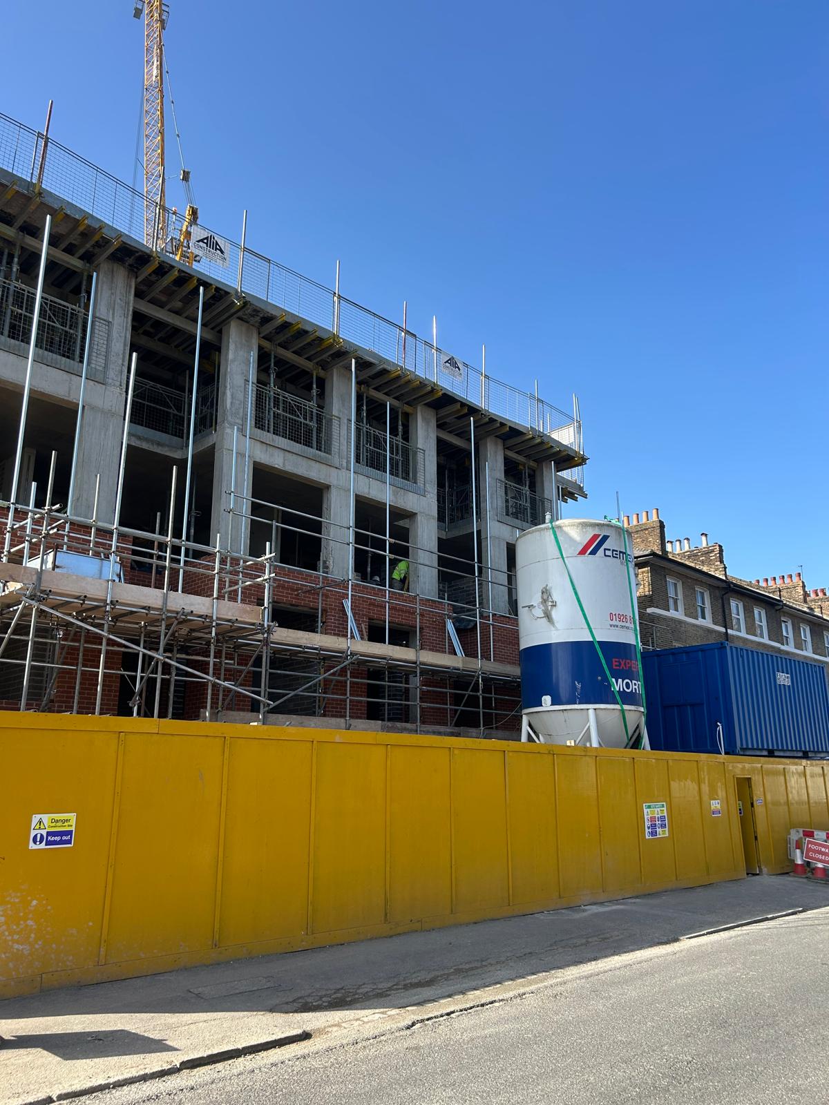 The site from the street, framed by the terraced houses behind the hoarding.